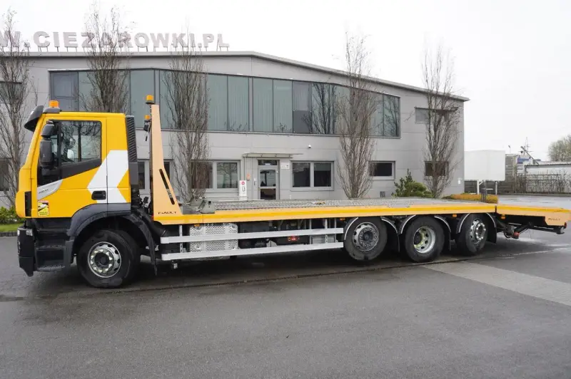 Żółty Iveco Stralis z platformą transportową stoi przed budynkiem.