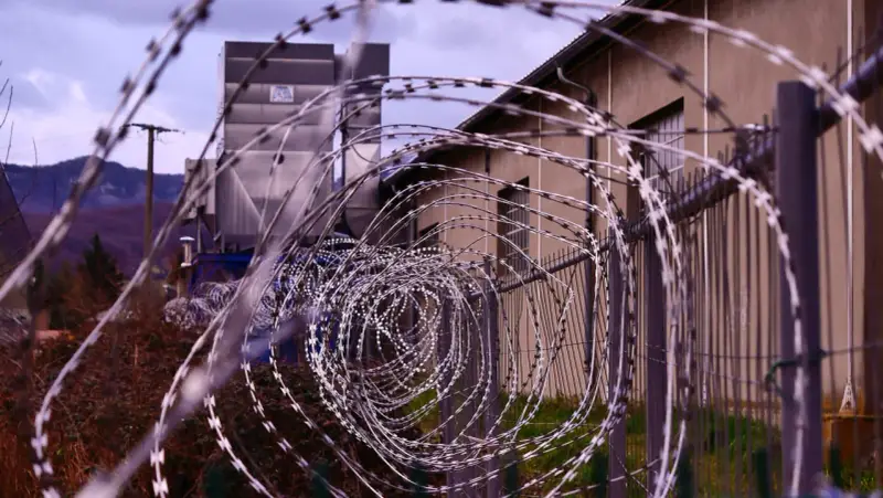 Kolczasty drut tworzy spiralę przed budynkiem. W tle widać góry i przemysłową konstrukcję. Może to miejsce, gdzie zapadały decyzje komisji penitencjarnej.