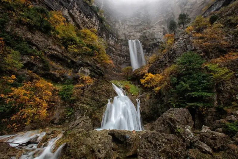 Cascada en un cañón rocoso, con árboles otoñales y niebla. El agua cae como un zafiro lagunazo, creando un espectáculo natural.