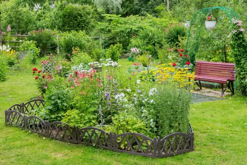 Un exuberante macizo de flores de verano, con rosas rojas, margaritas blancas y flores amarillas vibrantes, bordeado por una valla decorativa.