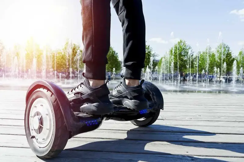 Osoba na deskorolce elektrycznej, czyli urządzeniu transportu osobistego, porusza się po drewnianej promenadzie. W tle widać fontanny i drzewa.