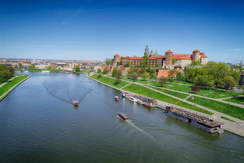 Widok na Wawel i Wisłę. Kilka łodzi pływa po rzece, a na brzegu zielony park i zabudowania.