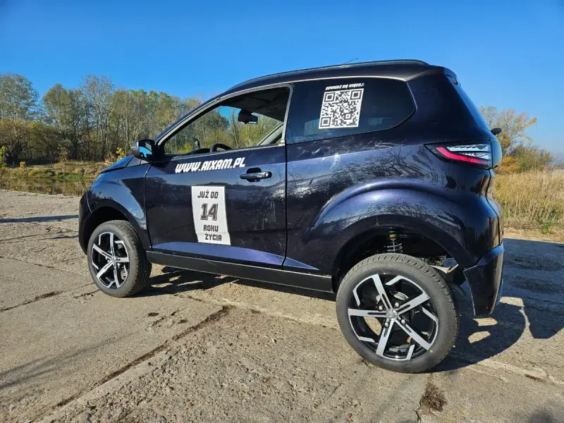 Ciemnoniebieskie Aixam auto z naklejkami "JUŻ OD 14 ROKU ŻYCIA" i kodem QR stoi na tle jesiennego krajobrazu.