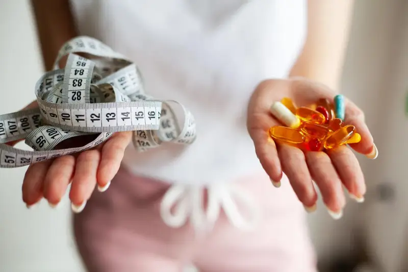 Dwie dłonie: jedna z miarką krawiecką, druga z kolorowymi kapsułkami – symbolizują wybór między dietą a środkami na odchudzanie.