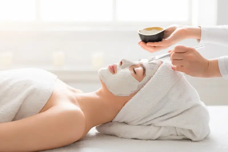 A spa technician applies a white mask to a woman's face. She's wrapped in a towel, enjoying a relaxing facial or massage first.