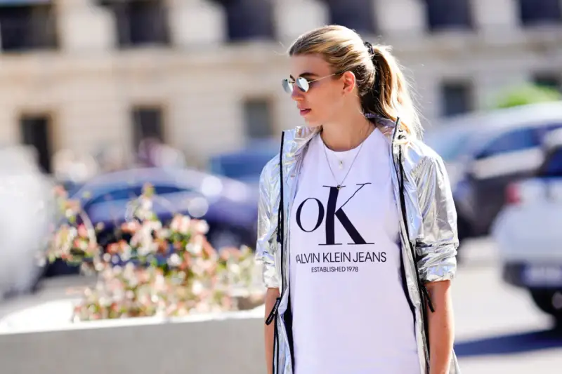Kobieta w srebrnej kurtce CK damskiej i białej koszulce z logo Calvin Klein Jeans.