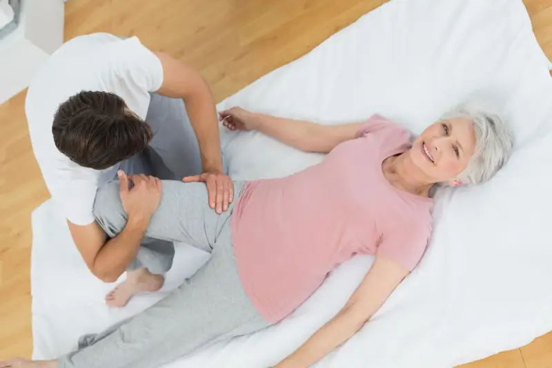 Physiotherapeut unterstützt ältere Frau bei Übungen für den Beckenboden. Sie lächelt zufrieden.