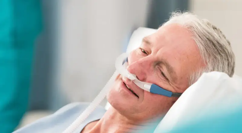 Älterer Mann mit Nasenkanüle, der entspannt im Bett liegt. Die Sauerstoffzufuhr mit **high flow** hilft ihm beim Atmen.