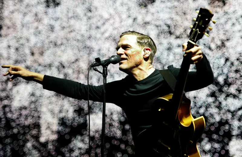 Bryan Adams w Polsce, śpiewa i gra na gitarze na tle świetlistej scenografii.
