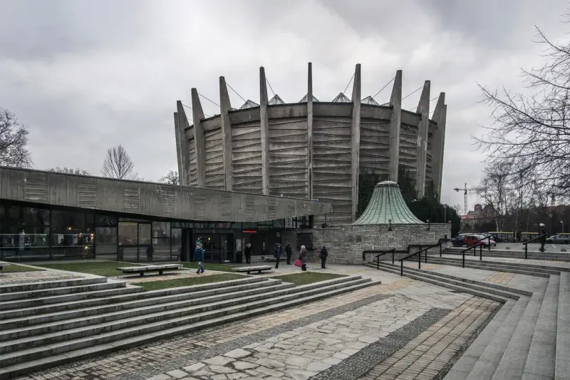 Monumentalny budynek w stylu brutalizm z betonowymi żebrami i szklaną fasadą. Schody prowadzą do wejścia, a wokół zielone trawniki.