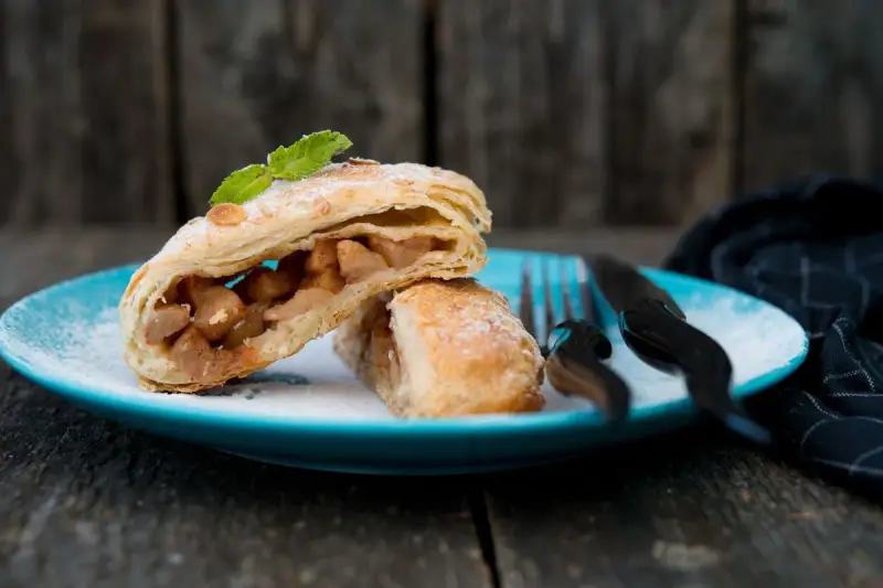 Delizioso strudel pere cioccolato e amaretti, con ripieno di mele e un tocco di menta, su un piatto blu cosparso di zucchero a velo.