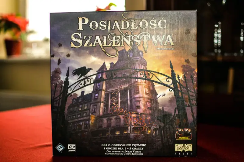 Pudełko gry "Posiadłość Szaleństwa" z mroczną posiadłością i wroną na bramie. Dodatki do tej gry planszowej zapowiadają nowe, przerażające przygody.