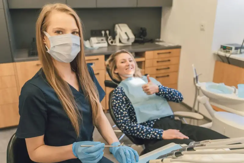Uśmiechnięta pacjentka daje kciuk w górę podczas pierwszej wizyty u dentysty po latach. Dentystka w masce i rękawiczkach przygotowuje narzędzia.