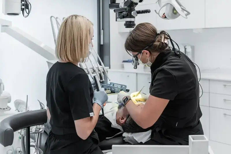 Dwie dentystki pracują przy pacjencie. Jedna używa mikroskopu, druga trzyma narzędzie. To obrazek pokazujący, jak zostać dentystą.