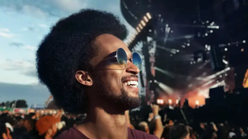 Un homme souriant avec des lunettes de soleil profite d'un concert en plein air. Il porte des bouchons d'oreilles, se demandant pourquoi pas de bouchon concert pour protéger son audition.