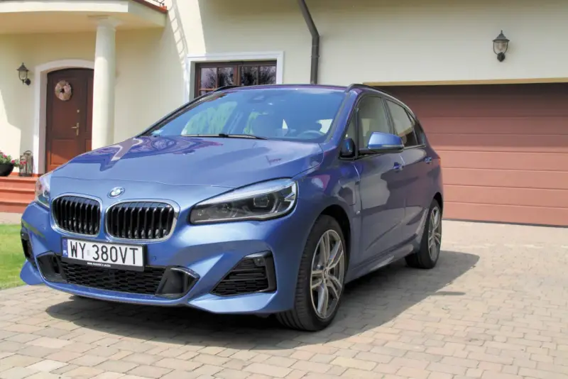 Niebieskie BMW 218i zaparkowane przed domem z brązową bramą garażową.