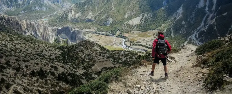 Widok na dolinę z góry. Turysta w czerwonej kurtce i plecaku zastanawia się, co zabrać w góry.