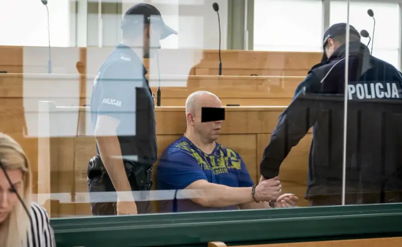 Skruszony gangster w kajdankach, prowadzony przez policjantów w sali sądowej.