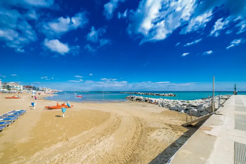 Spiaggia di Bellaria Mare con lettini, bagnini arancioni e mare azzurro sotto un cielo blu con nuvole bianche.