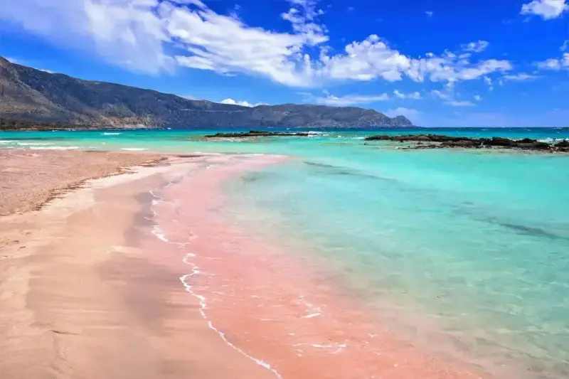 Różowy piasek plaży Elafonisi w Grecji, turkusowa woda i góry w tle.