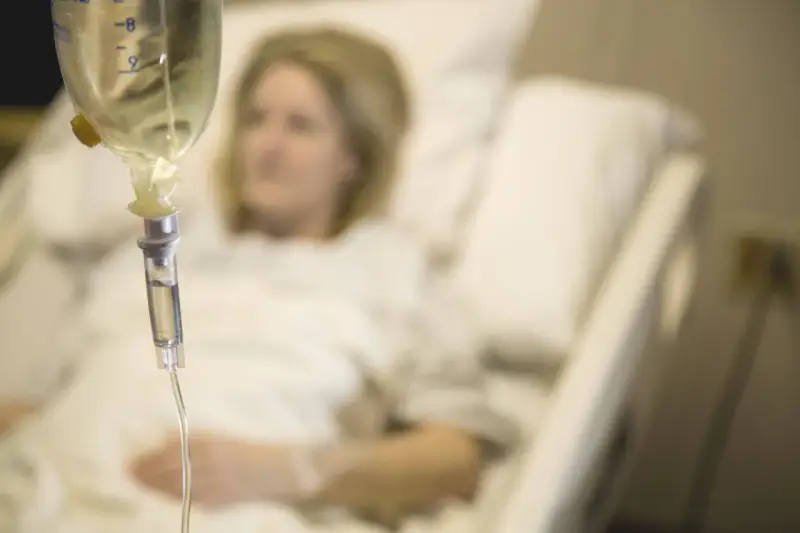 Infusion vorbereiten: Eine Frau liegt im Krankenhausbett, während eine Infusionstropfflasche und ein Tropfschlauch im Vordergrund zu sehen sind.