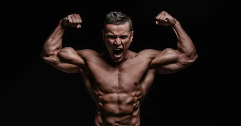 Mężczyzna z wyrzeźbionymi mięśniami napina bicepsy, krzycząc z wysiłku. Jego sylwetka sugeruje intensywny trening, być może z użyciem clenbuterolu.