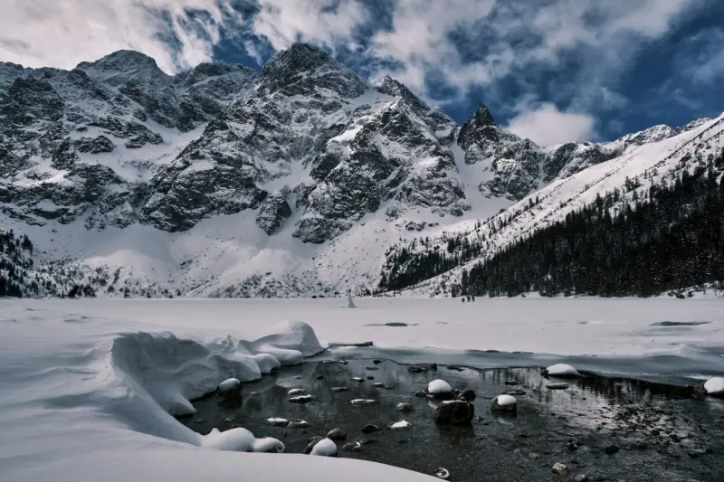 Zamarznięte Morskie Oko, otoczone ośnieżonymi szczytami Tatr. W oddali widać ludzi spacerujących po lodzie, a na pierwszym planie płynie strumyk.
