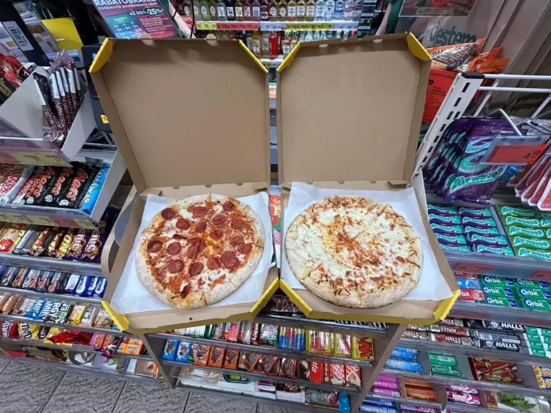 Dwie pizze w pudełkach, jedna z salami, druga serowa. Obok półki ze słodyczami i napojami w sklepie Żabka.