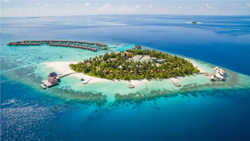 Rajskie Malediwy wiza: wyspa z bungalowami na wodzie, plażą i palmami, otoczona turkusową wodą.