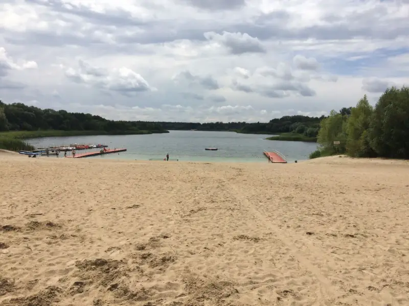 Piaskowa plaża nad jeziorem, z pomostami i łódkami. W oddali las, chmury na niebie. Idealne miejsce na wypoczynek nad wodą.