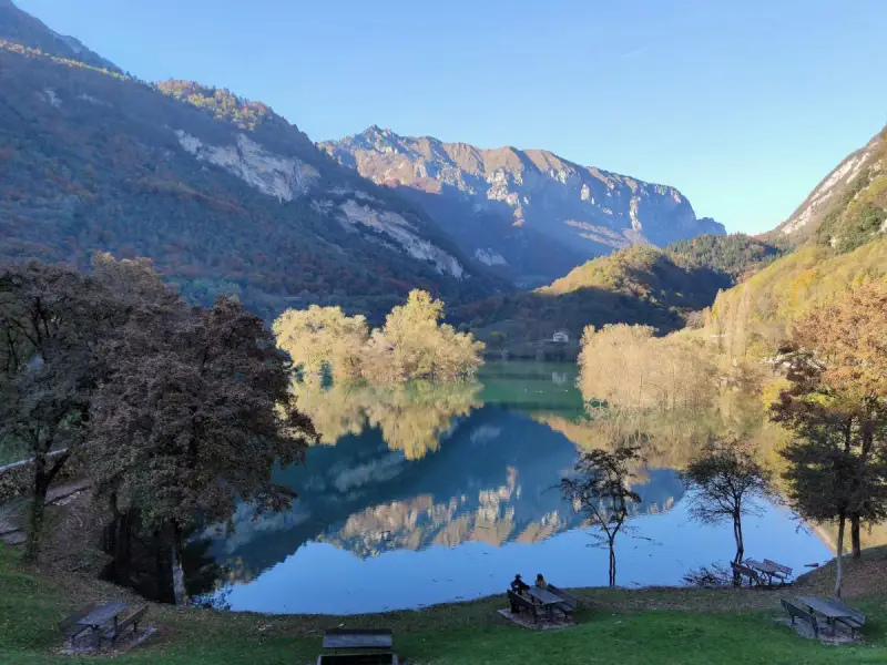 Il lago di Tenno riflette montagne imponenti e alberi autunnali. Due persone siedono su una panchina, godendosi la quiete.