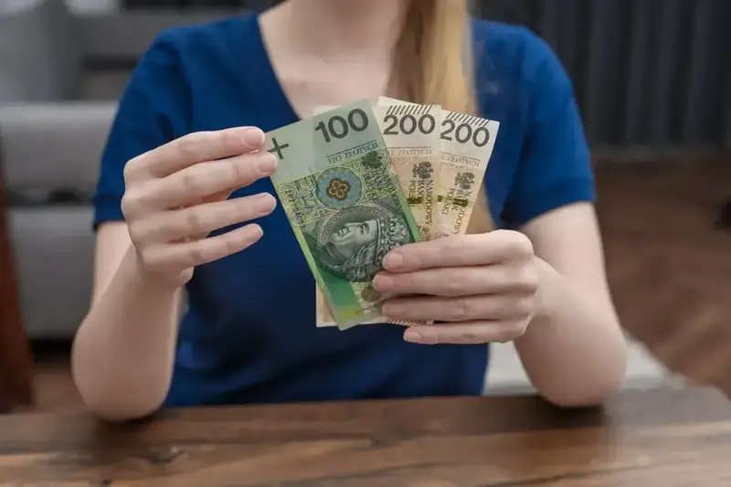 Dłonie trzymają polskie banknoty: 100 zł i dwa razy po 200 zł. To może być część zasiłku rodzicielskiego.