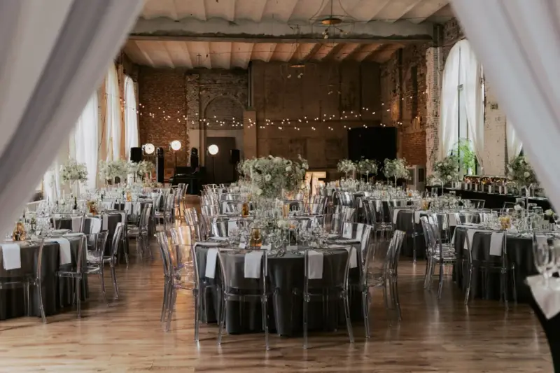 Eleganckie wesele w industrialnym stylu. Stoły nakryte szarymi obrusami, ozdobione białymi kwiatami i transparentnymi krzesłami. Girlandy świateł tworzą magiczny nastrój.