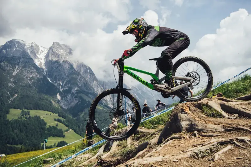 Troy Brosnan in mid-air on a green downhill bike, with a dramatic mountain backdrop.