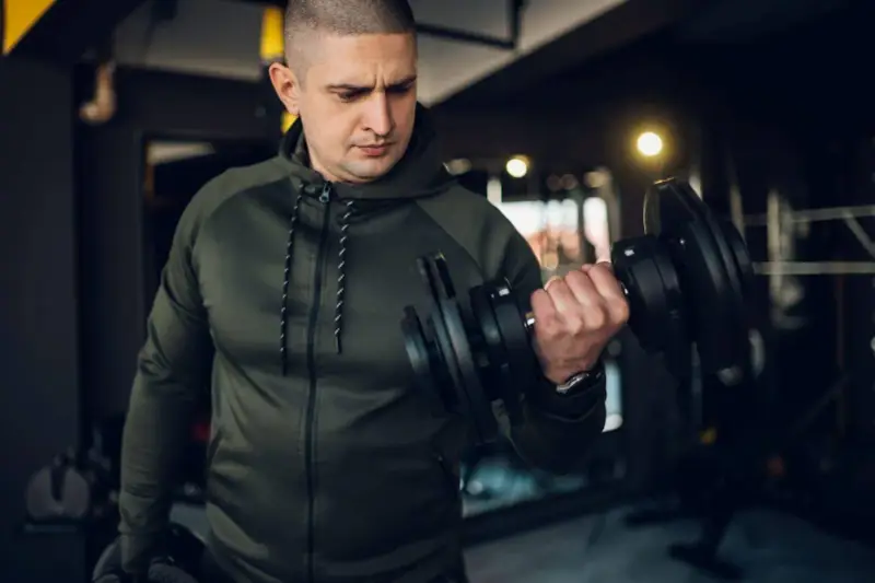 Mężczyzna w zielonej bluzie damskiej na siłownię ćwiczy biceps z hantlem.
