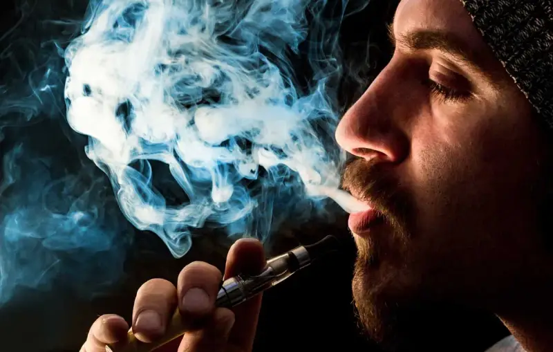 Hombre vapeando, el vaper sin nicotina es malo. El humo azul forma figuras fantasmagóricas.
