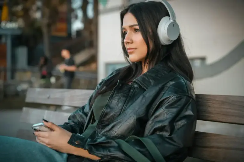 Młoda kobieta w słuchawkach, słuchająca podcastu o wymowie francuskiej, siedzi na ławce, trzymając telefon.