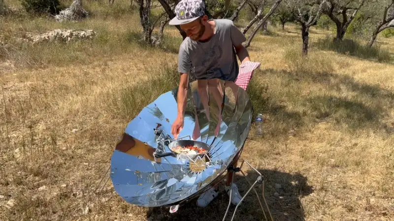 Un homme cuisine avec un four solaire parabolique, démontrant le fonctionnement de cet appareil écologique en plein air.