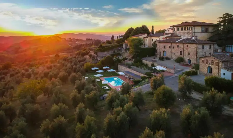 Un tramonto dorato illumina un agriturismo con piscina, immerso tra ulivi e colline, perfetto per un tour della Toscana.