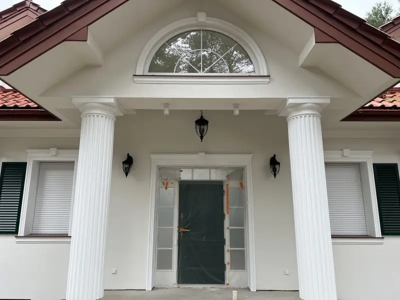 Elegancki pilaster zdobi fasadę domu, wspierając dach nad wejściem. Obok okna i drzwi, tworzy klasyczną kompozycję.