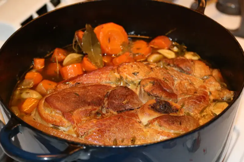 Rouelle de porc mijotant dans une cocotte avec des carottes, de l'ail et des herbes.