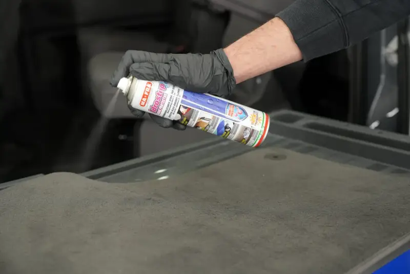 Mano con guanto nero spruzza spray Idrostop su tessuto grigio per impermeabilizzare.