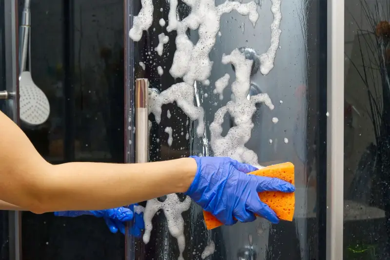 Main bleue, éponge orange pour nettoyer paroi douche. Mousse et bulles sur la paroi vitrée.