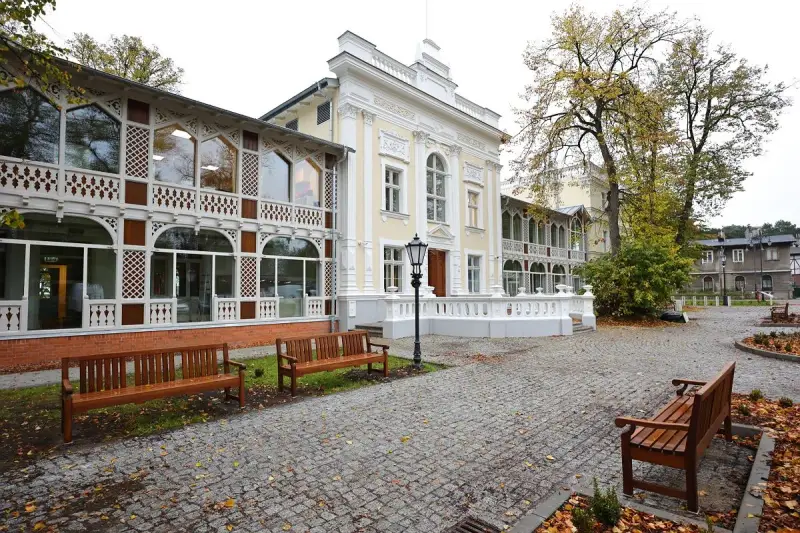 Elegancki budynek z ażurowymi werandami, przypominający dawny **Hevelianum dom zdrojowy**. Brukowany dziedziniec z ławkami i jesiennymi liśćmi.