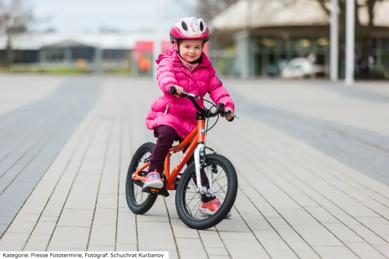 Ein fröhliches Mädchen in pinker Jacke und Helm sitzt auf einem orangefarbenen Kinderfahrrad. Die Frage "kinderfahrrad 4 jahre wieviel zoll" wird durch die Größe des Fahrrads beantwortet.
