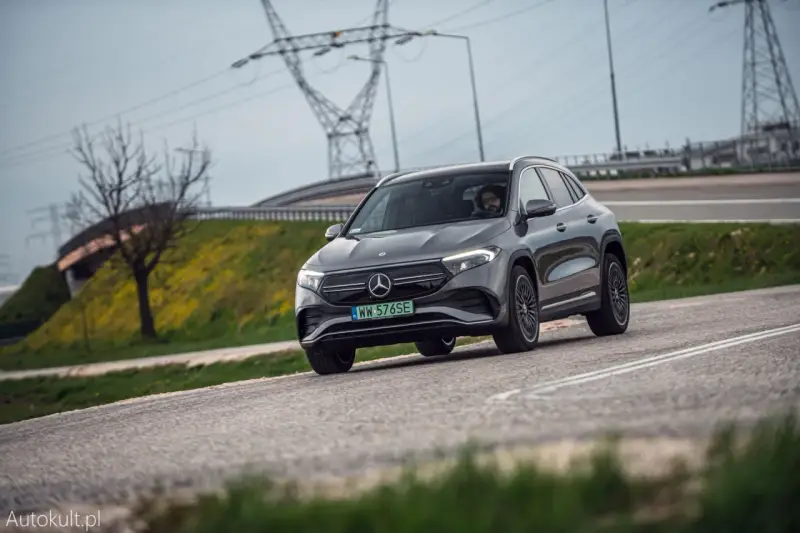Szary Mercedes-Benz EQA jedzie po drodze, w tle linie energetyczne i drzewo.