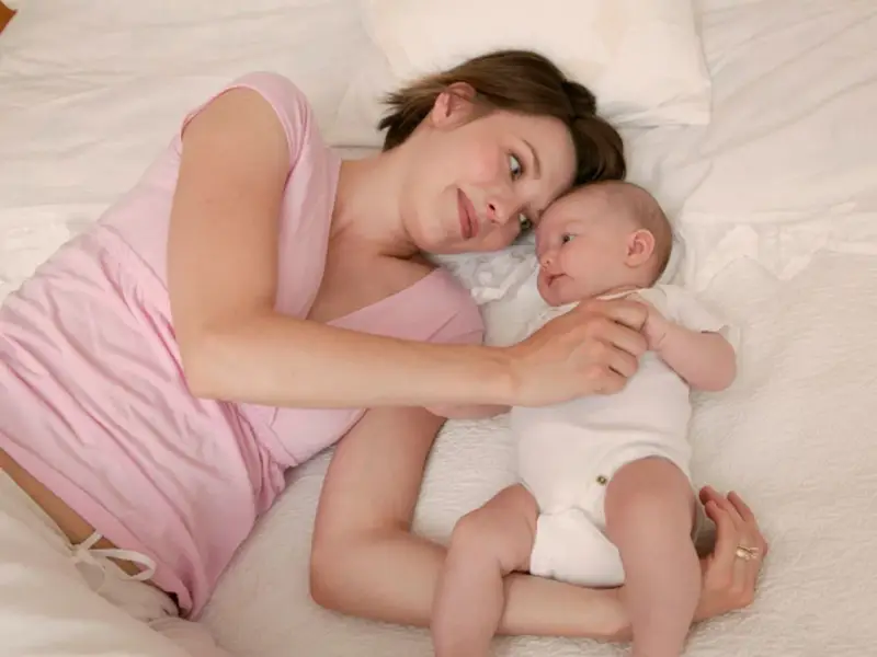 Mama patrzy z miłością na swoje 5 tygodniowe dziecko leżące obok niej na łóżku.
