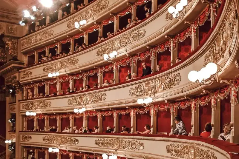 Widownia La Scali, pełna widzów w lożach, oczekuje na rozpoczęcie spektaklu. Bogaty repertuar teatru przyciąga miłośników sztuki.