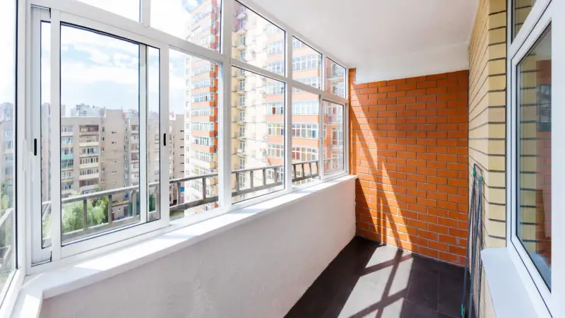 Wnętrze loggii z widokiem na miejską zabudowę. Ściany z cegły i tynku, przeszklony balkon z białymi ramami okiennymi.