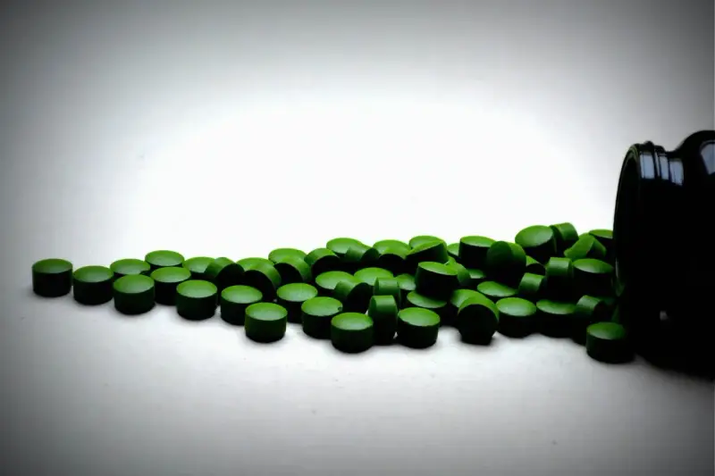 Zielone tabletki chlorelli rozsypane z czarnego pojemnika. Kiedy je brać: chlorella rano czy wieczorem?
